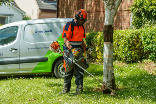 Image of a Greatwell Places operative dressed in PPE, strimming grass around a tree. A Greatwell Homes van is in the background.