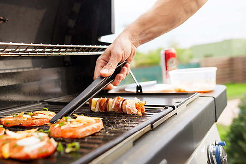 Hand cooking food on a barbecue grill with tongs. In the background there is a sunny garden.