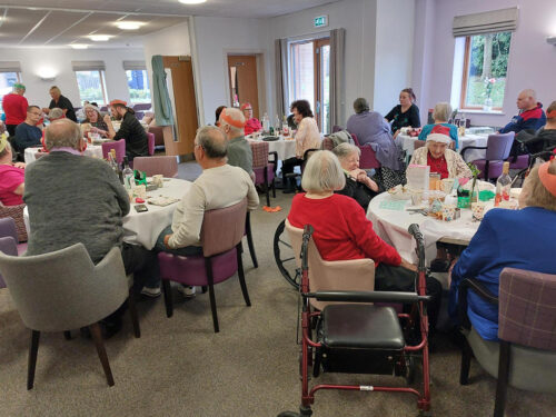 Photo of the communal room at Knights Court Independent Living scheme, full of customers sitting around round tables enjoying Christmas dinners and drinks. They are smiling and talking with each-other.